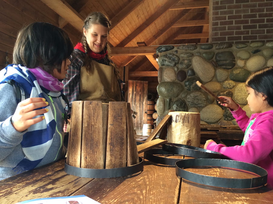 Two young girls learning to craft barrels from a period costumed interpreter at Fort Langley.